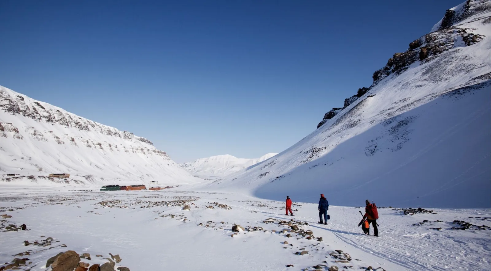 Three walkers crossing a snow-covered Svalbard valley under a deep blue sky