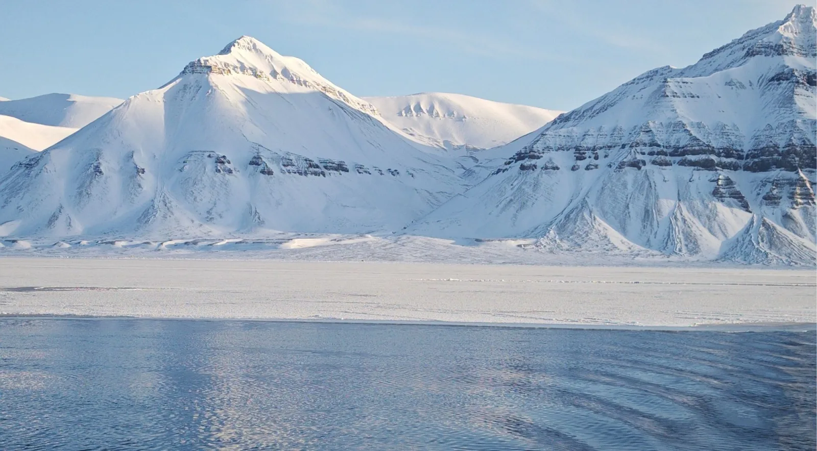 Snow-covered Svalbard mountains above a frozen shoreline and open fjord water