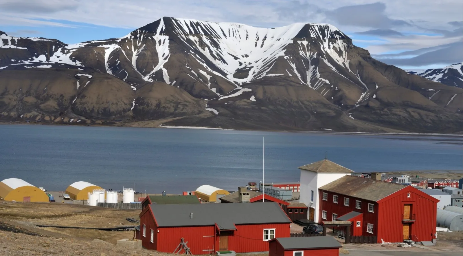 Red timber buildings on the shore of a Svalbard fjord below a striped snow-streaked mountain