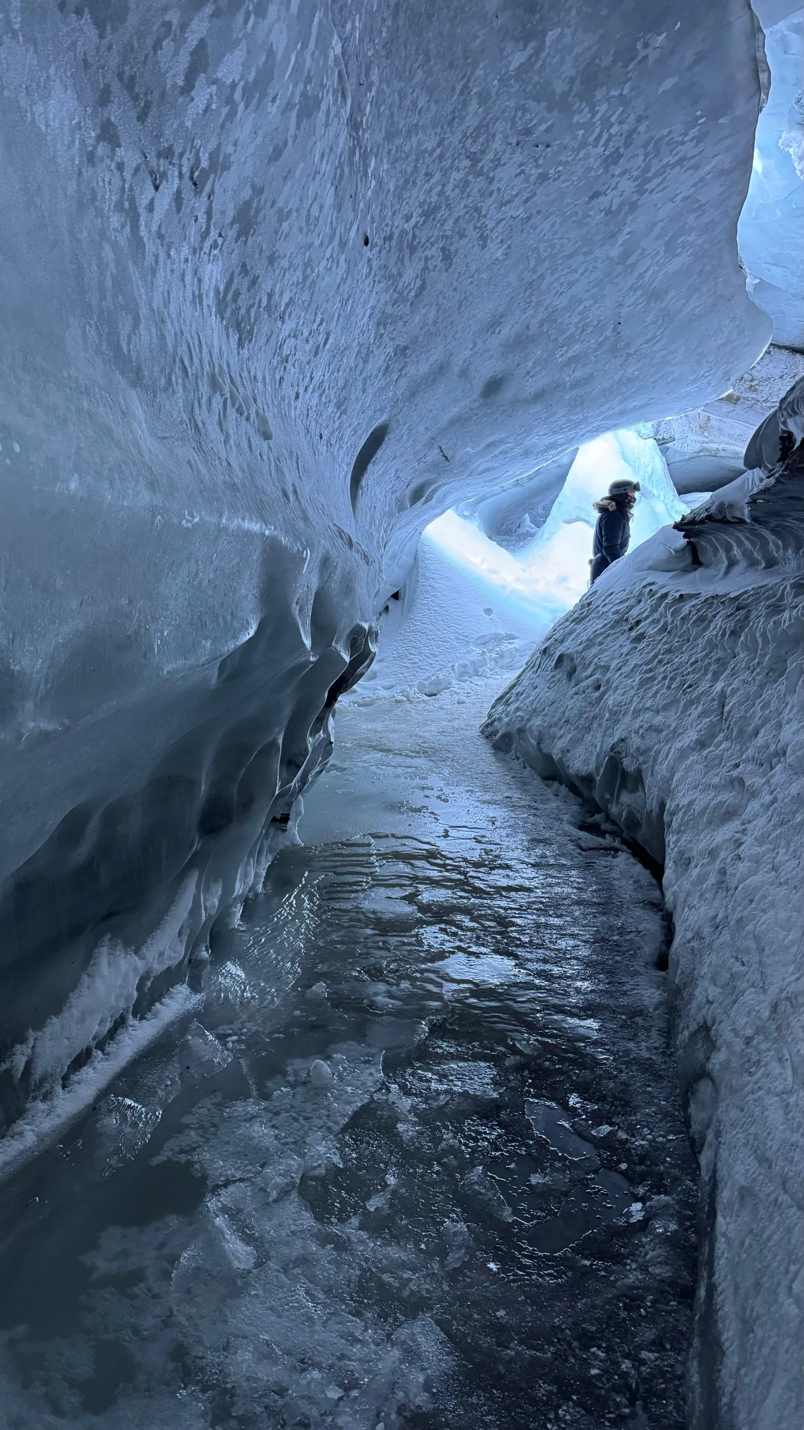 A visitor at the mouth of an ice cave, the meltwater channel running away into the glacier
