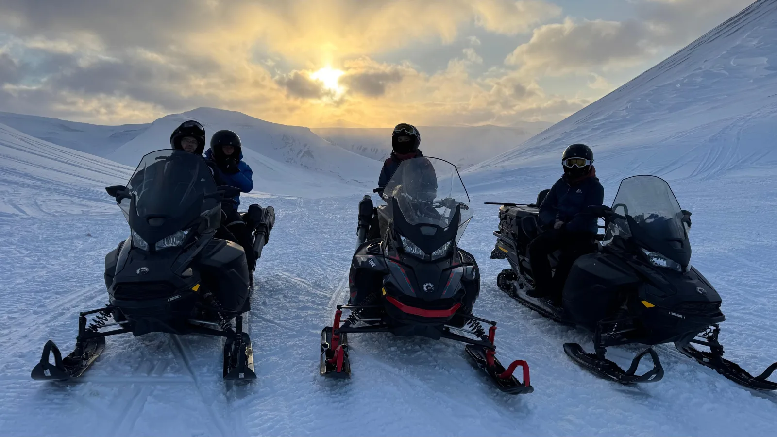 Three snowmobiles lined up on a snow slope at sunset with riders in helmets