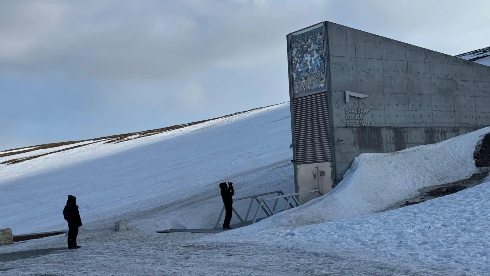 Two visitors photographing the Seed Vault's concrete wedge across a snow slope