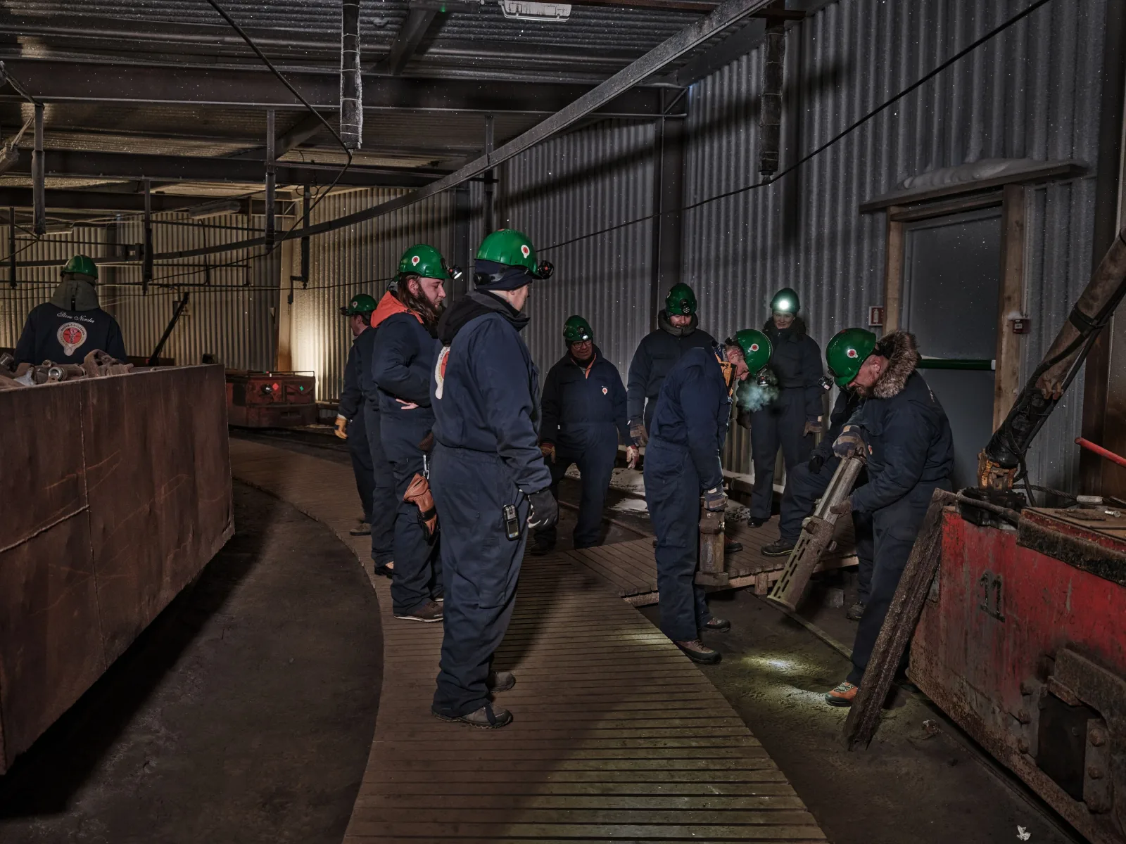 Group in green helmets and navy overalls being briefed beside a pneumatic drill in the mine workshop