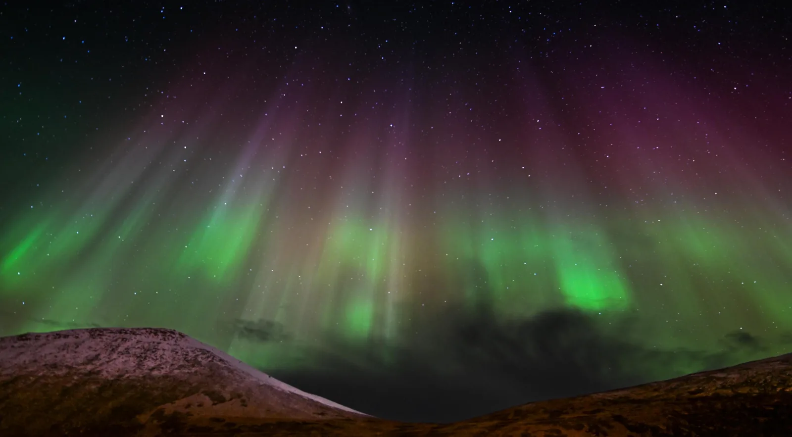 Curtains of green and magenta aurora falling behind a low snow-dusted ridge