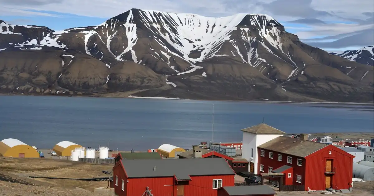 Red timber buildings on the shore of a Svalbard fjord below a striped snow-streaked mountain