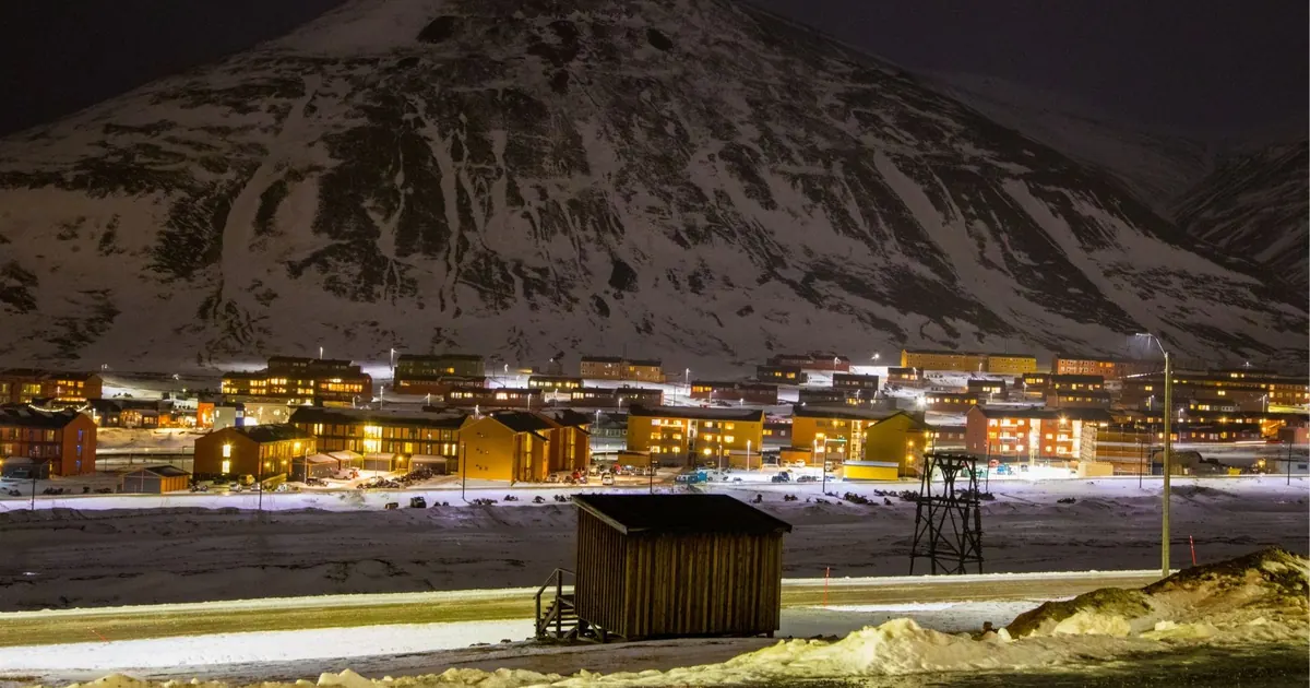 Longyearbyen lit up at night below a dark mountain, seen across the snow-covered valley floor