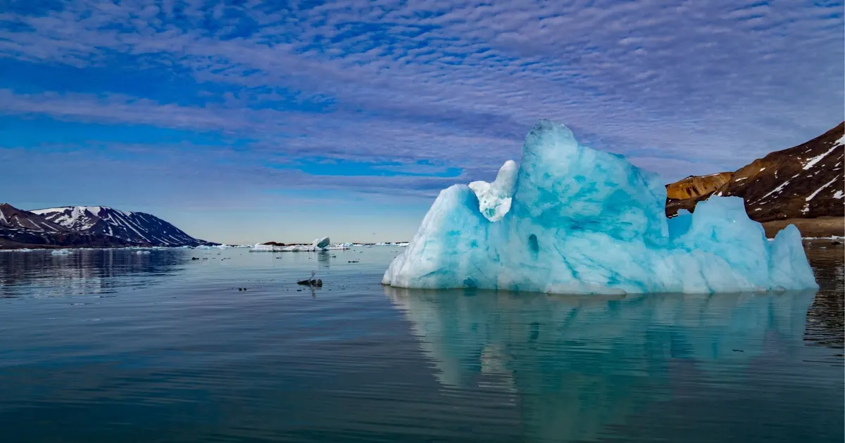 A blue iceberg floating on calm water in a Svalbard fjord beneath a streaked sky