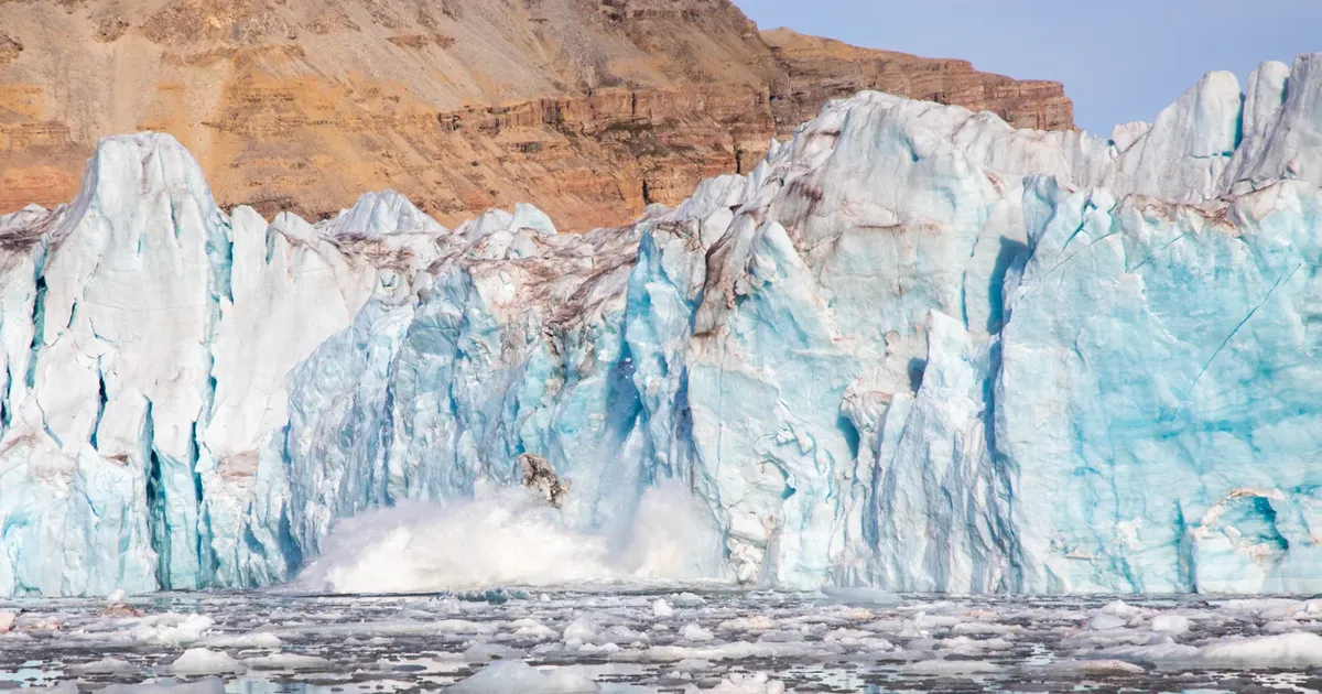 Blue-white glacier front calving into an ice-strewn fjord below red-brown Svalbard cliffs