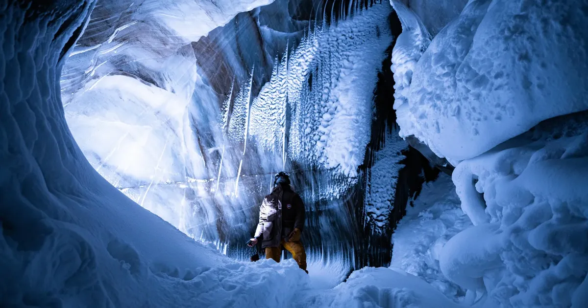 A person standing inside a Svalbard glacier ice cave with blue-lit ice walls curving overhead