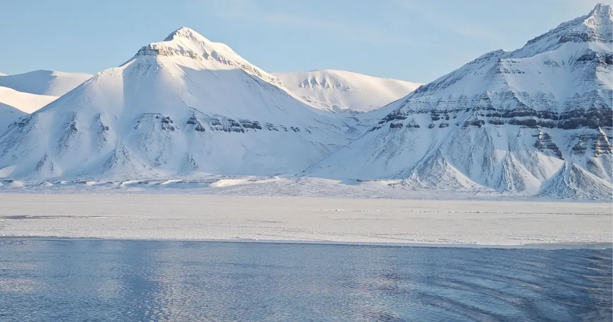 Snow-covered Svalbard mountains above a frozen shoreline and open fjord water in spring sunshine