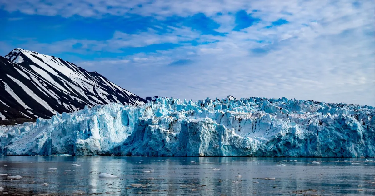 A pale blue glacier front rising from calm fjord water under a blue sky in Svalbard