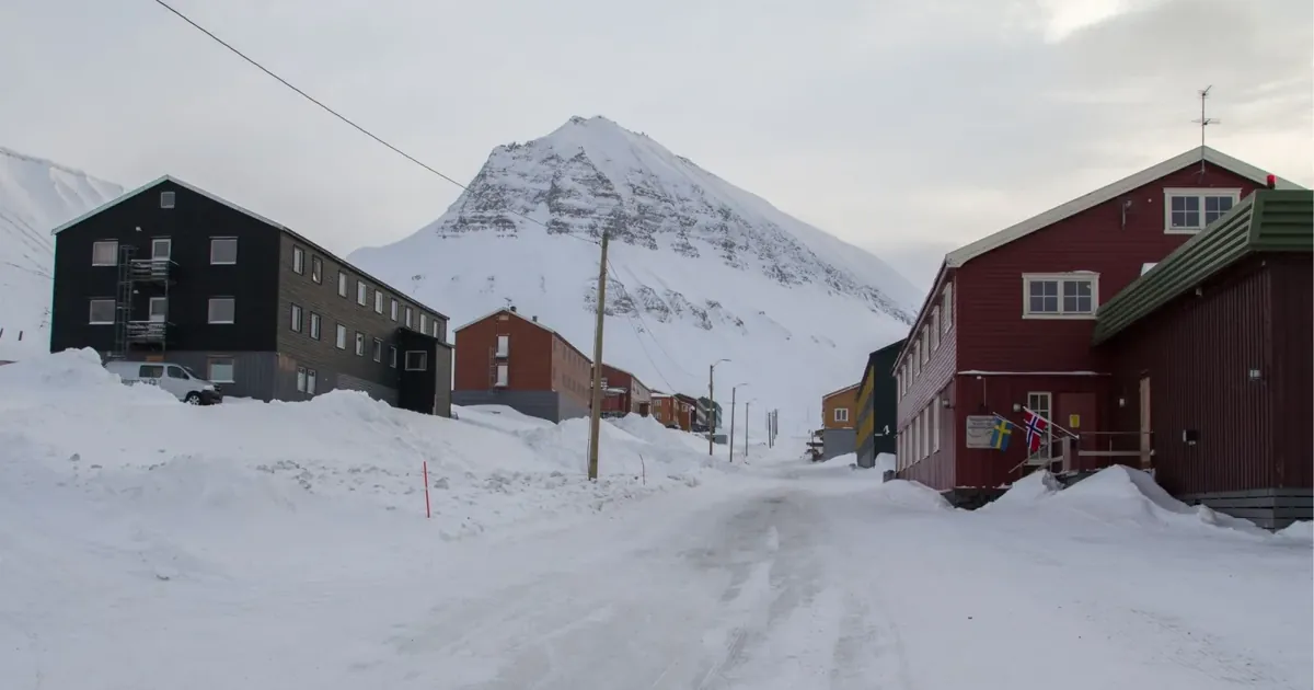 A snow-banked Longyearbyen street between red and black timber buildings, with a snow-covered peak at the end